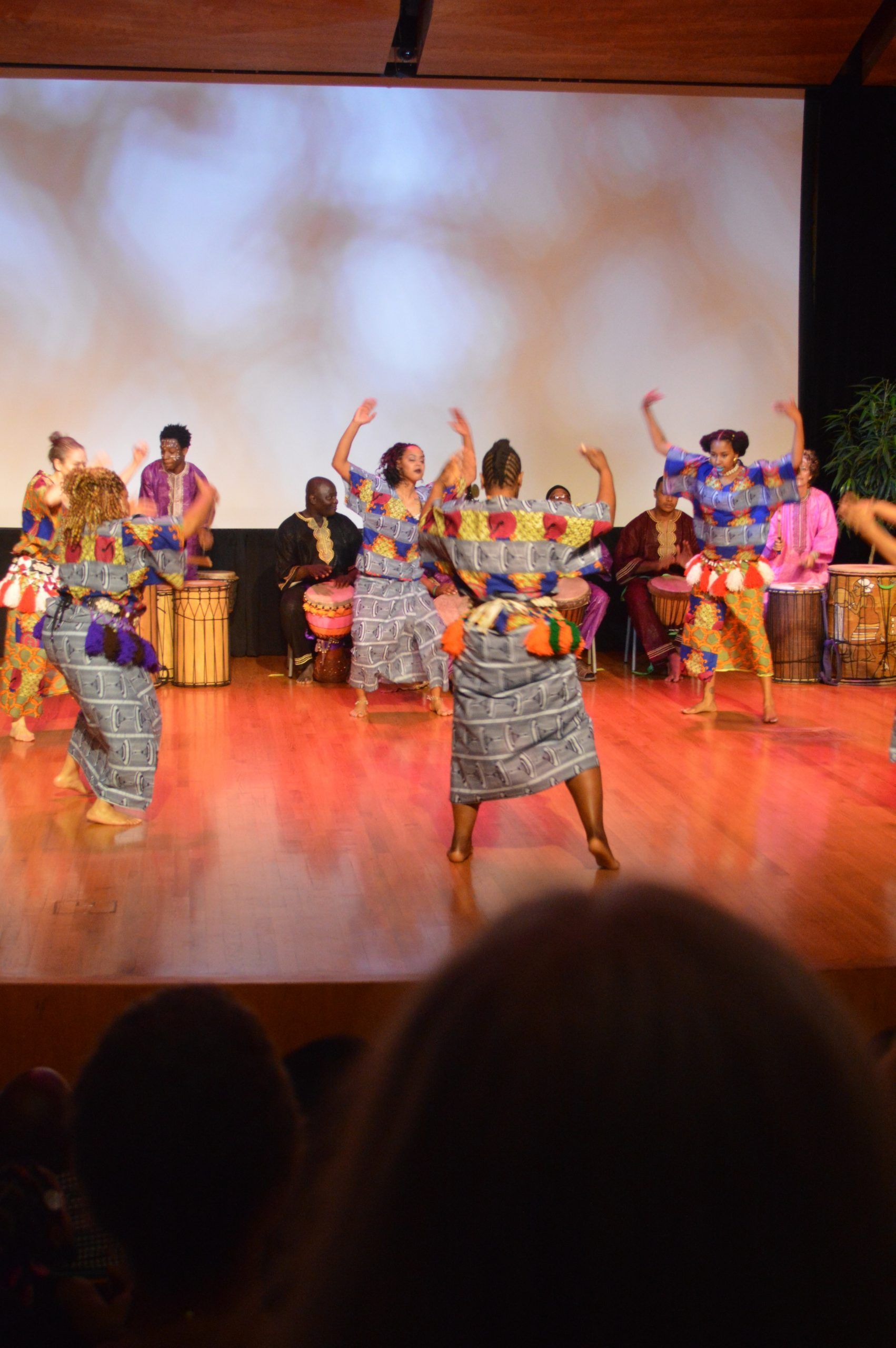 Dancers in colorful African attire perform on stage; drummers in the background.