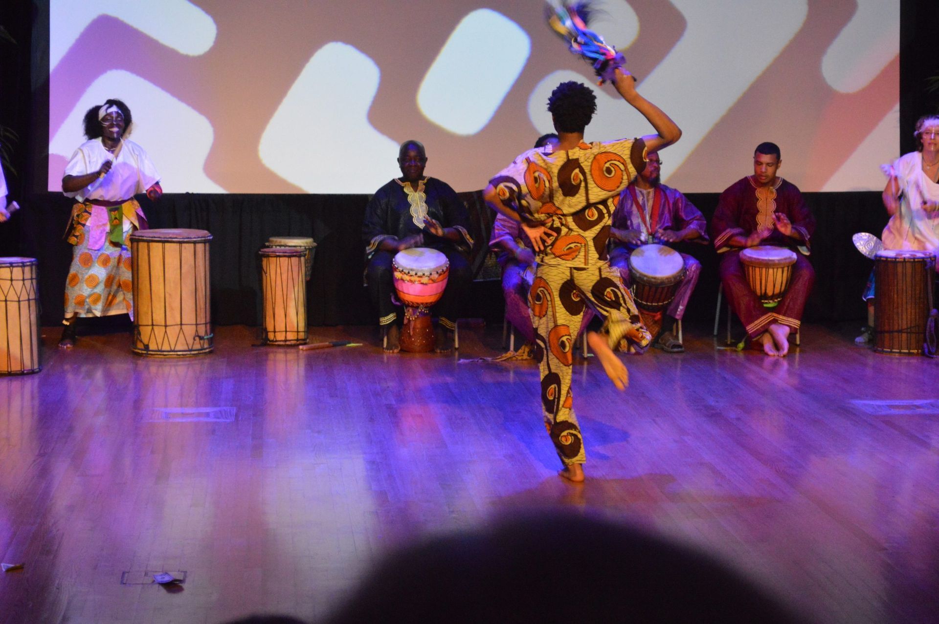 African dancers and drummers on stage, colorful costumes, performing with drums in front of a projected backdrop.