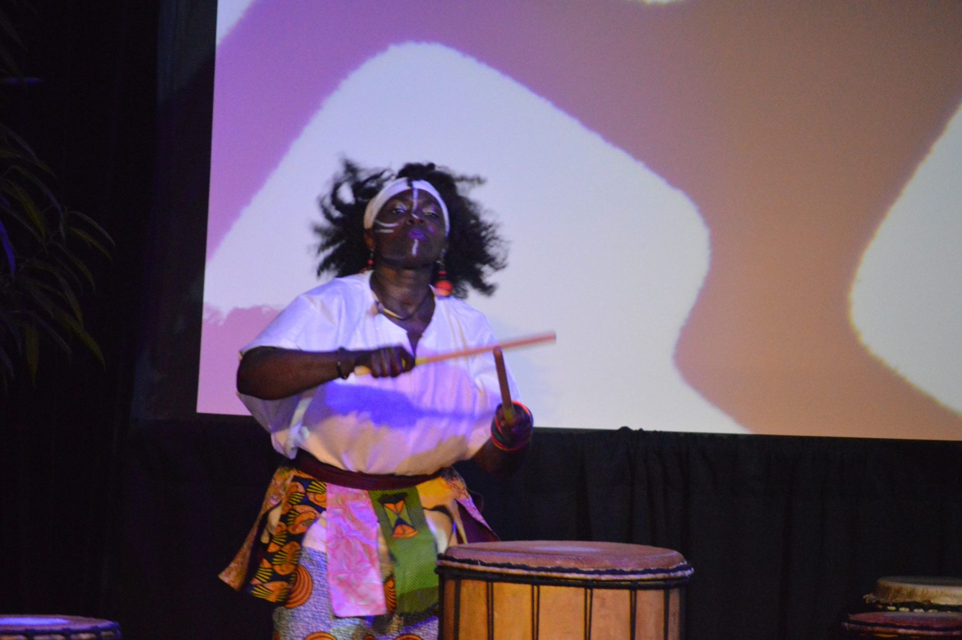 Woman playing a drum onstage, wearing white shirt, colorful skirt, face paint.