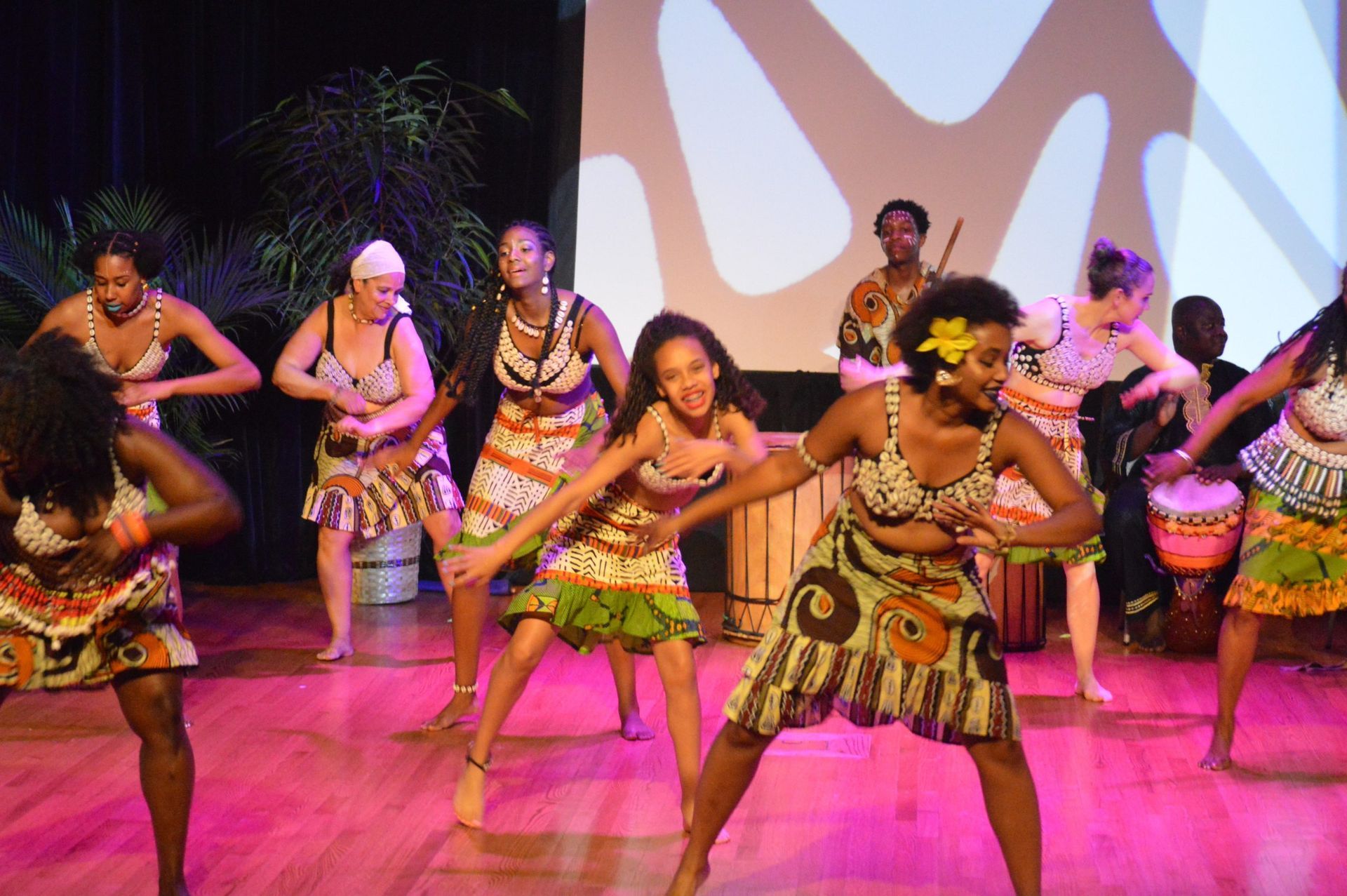 African dancers in colorful attire perform on a stage with musicians and vibrant background.