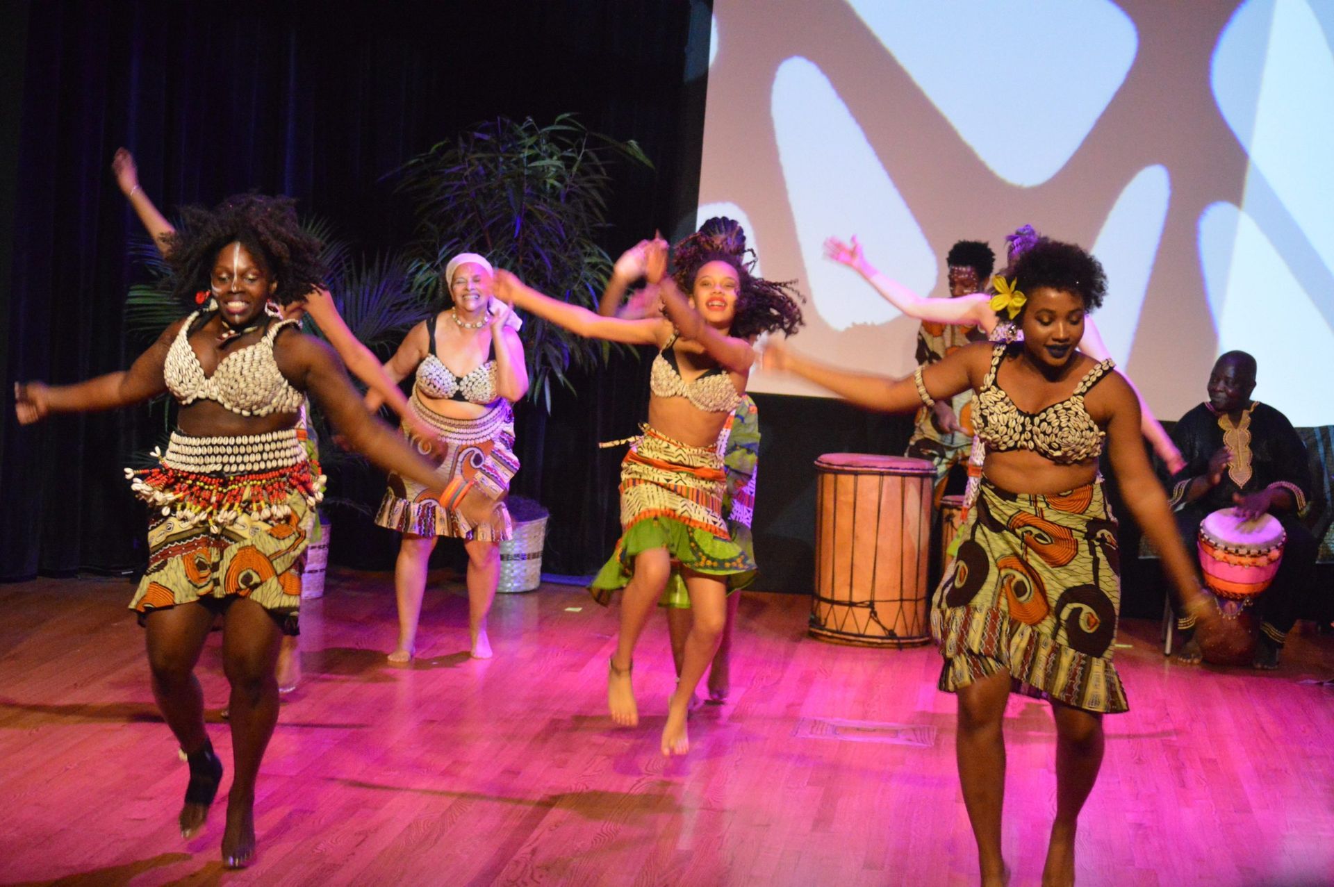 Dancers in African print attire perform on stage, arms outstretched, with drummers in the background.