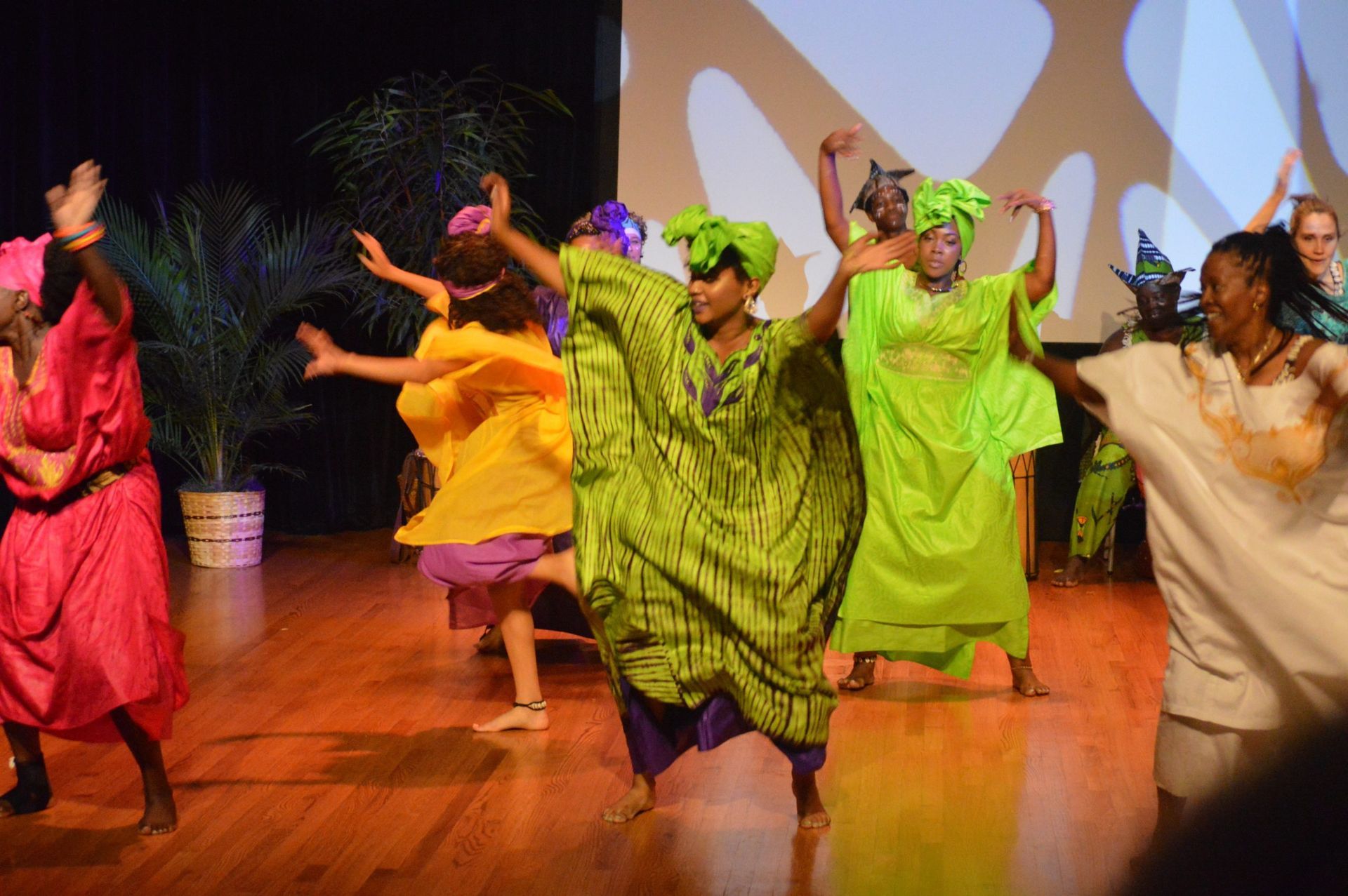 Dancers in colorful robes on stage, some with head wraps, performing a traditional dance.
