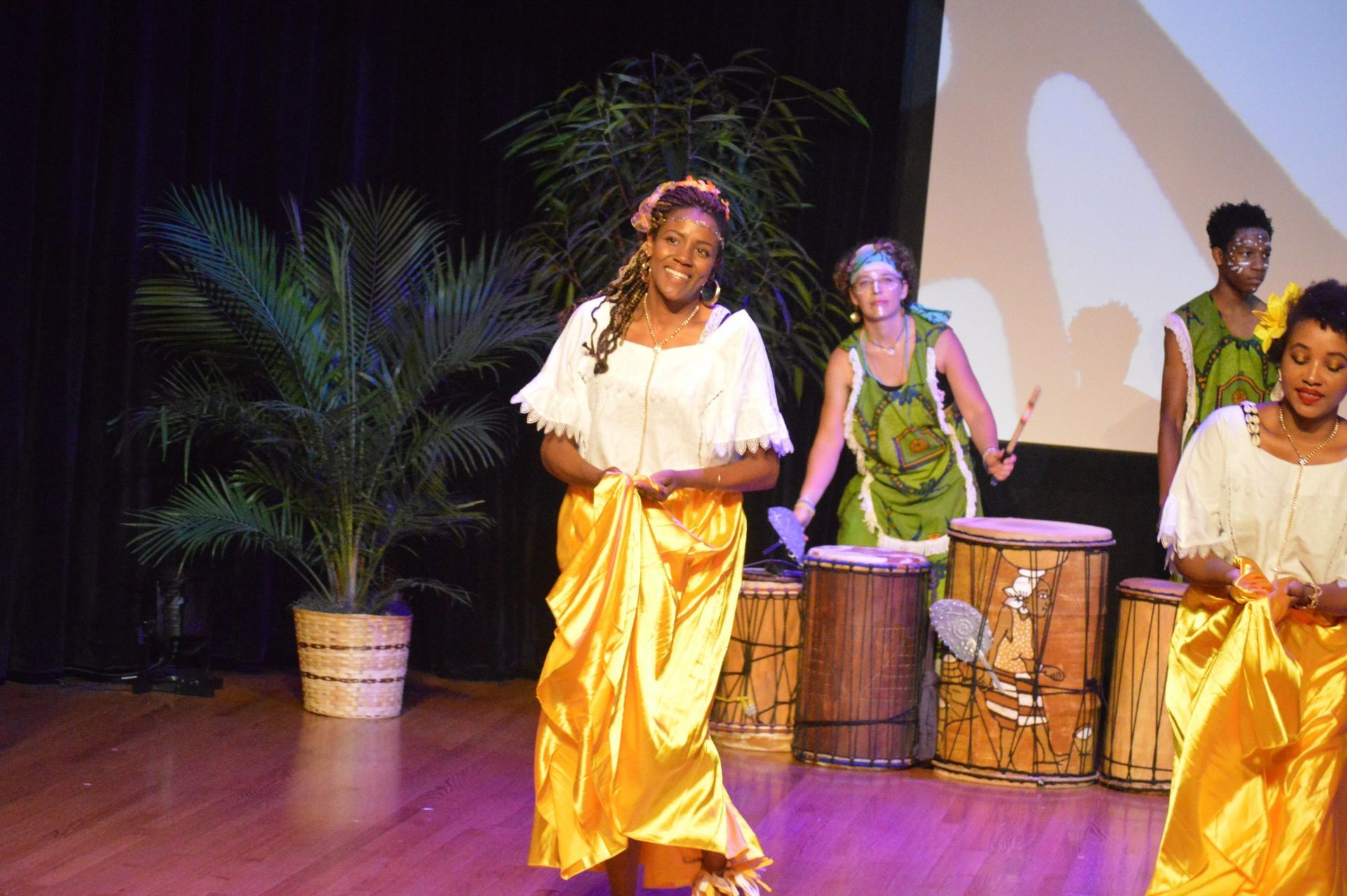 Woman in gold skirt and white top performs on stage with drummers.
