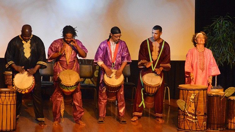 Five people playing drums on a stage, dressed in colorful traditional garments.