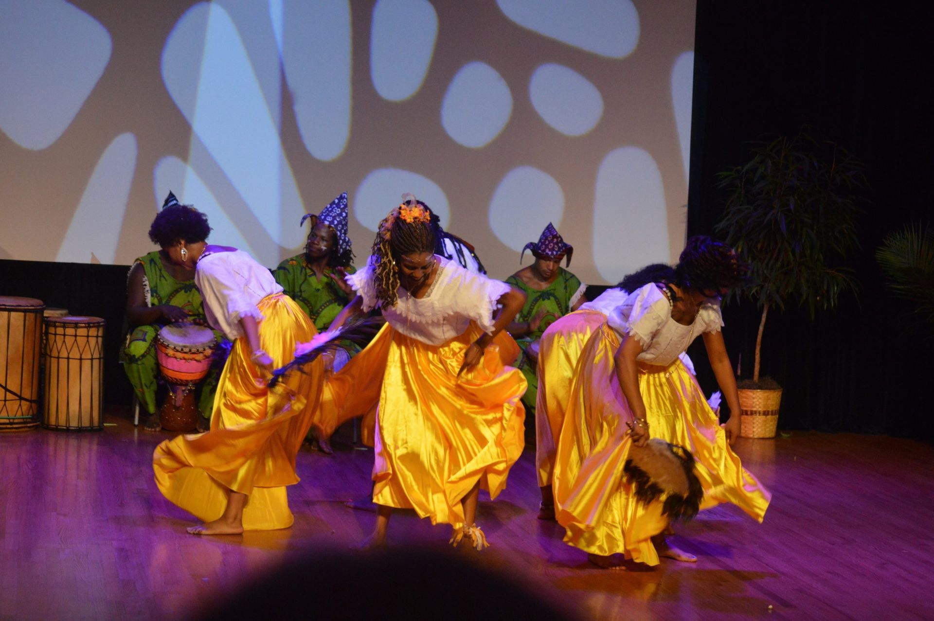 Dancers in white tops, yellow skirts, perform on stage with drummers; stage lit with abstract shapes, wooden drums.