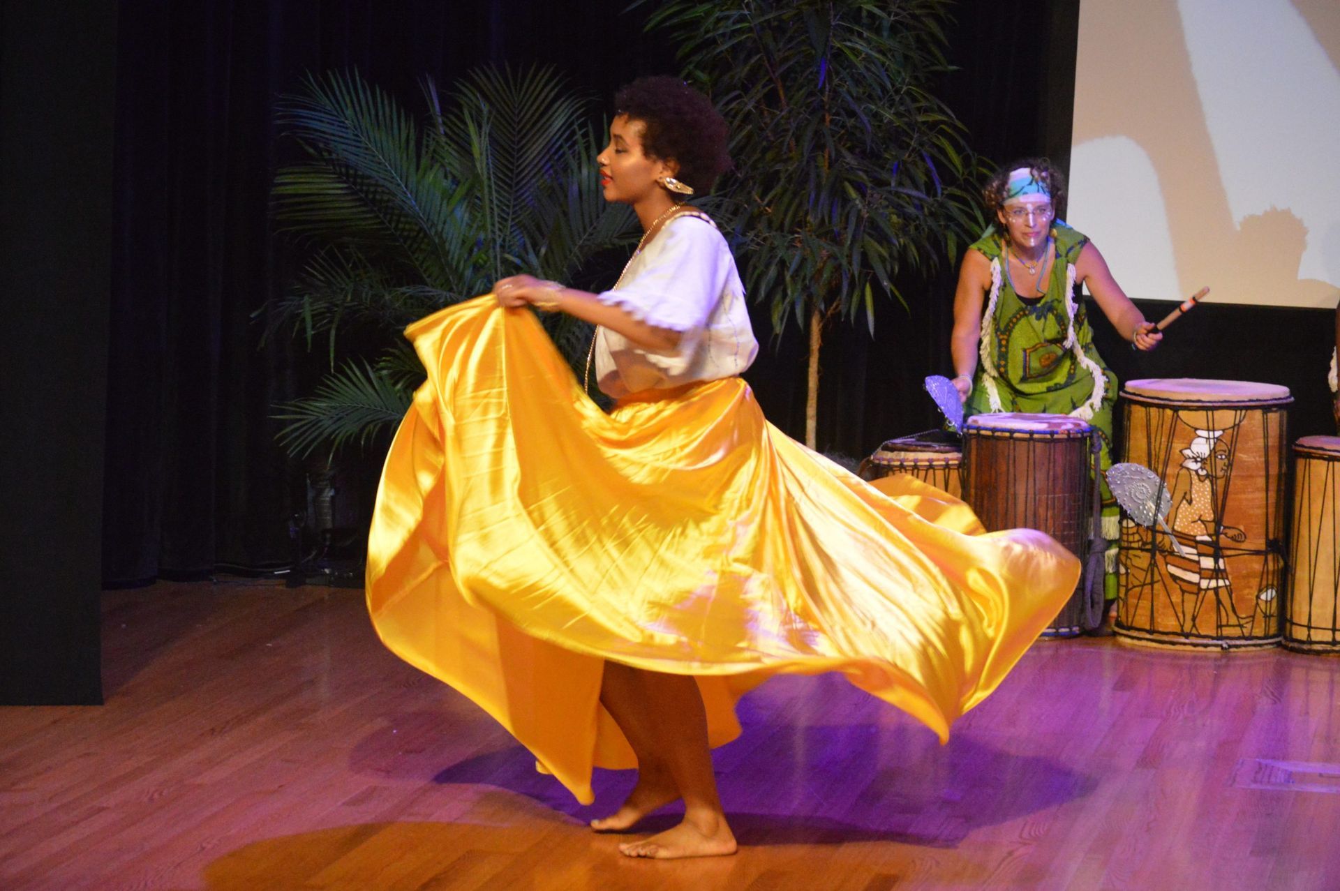 Woman in gold skirt dancing, drummer in background. Stage, tropical plants, and drums present.