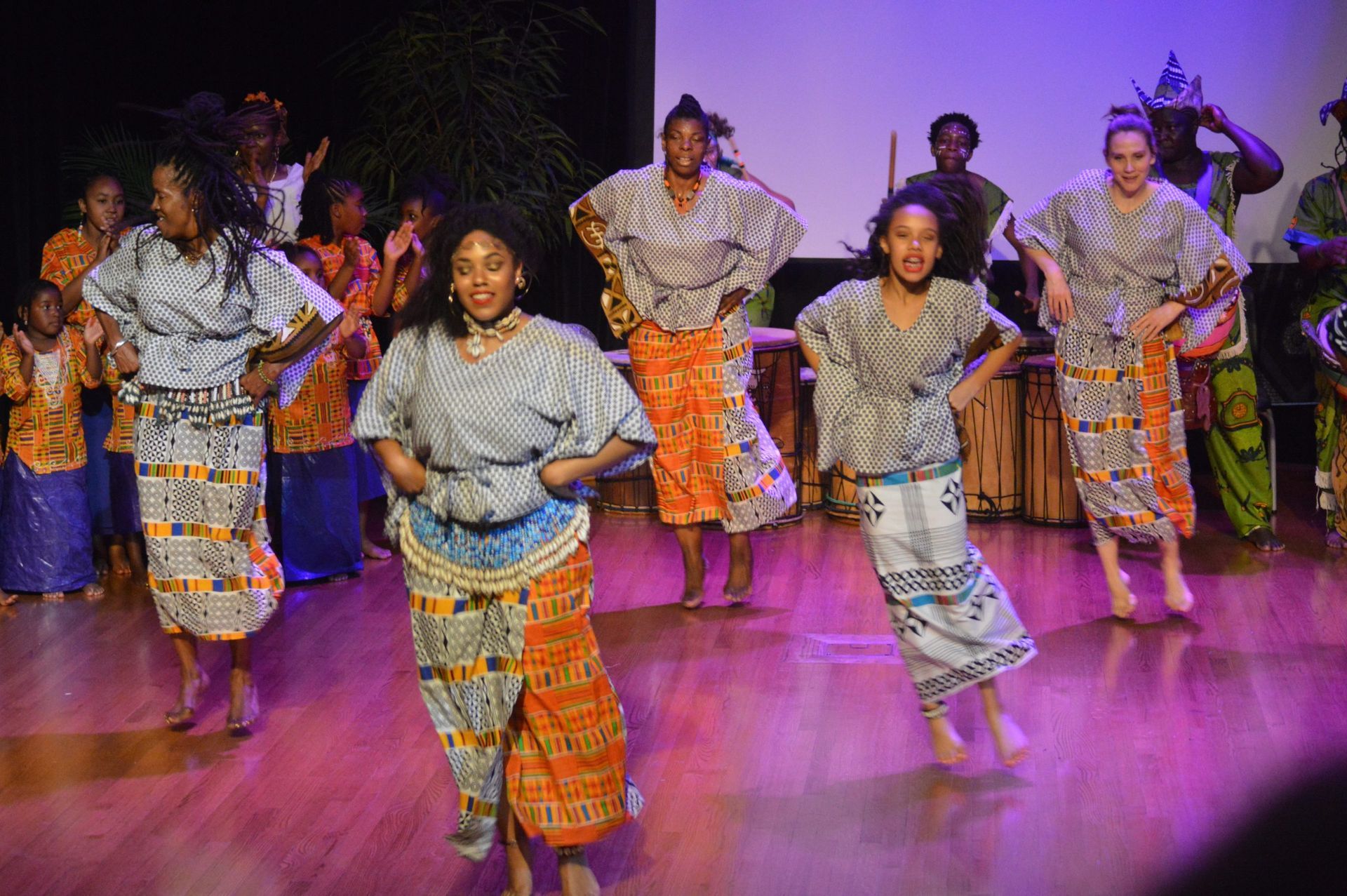 Dancers in patterned outfits perform on stage, hands on hips. Wooden drums and musicians are visible in the background.