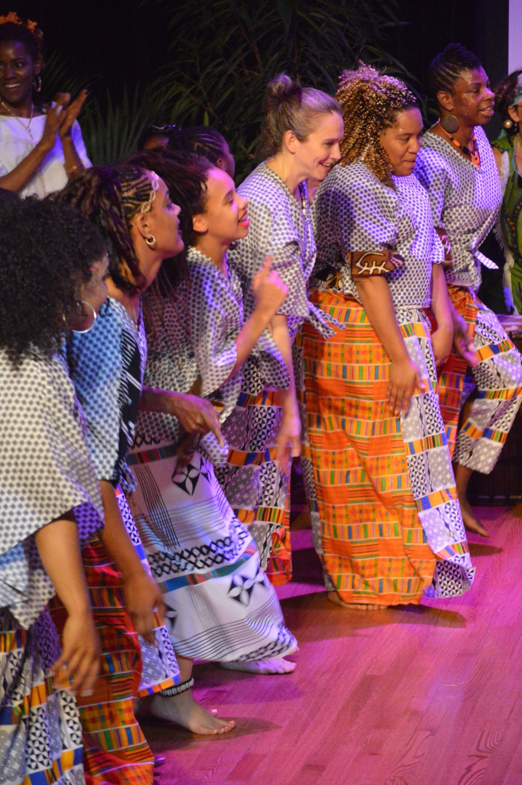 Women in patterned dresses dance on a stage with pink lighting.
