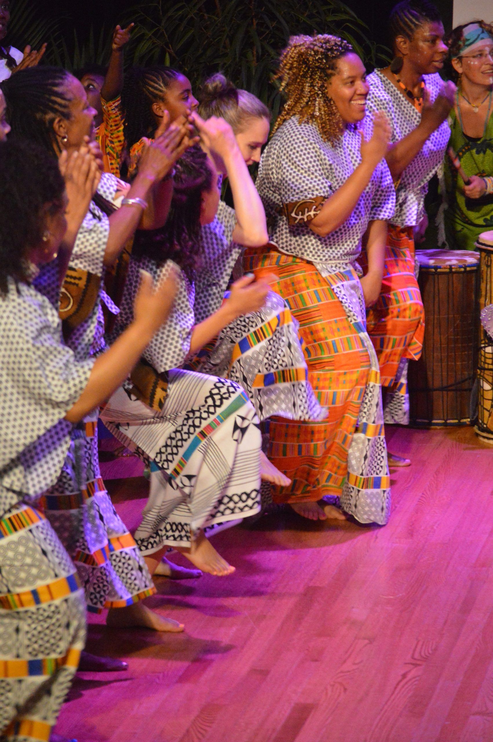 Group of people dancing, clapping, and smiling in traditional African clothing; indoor setting.