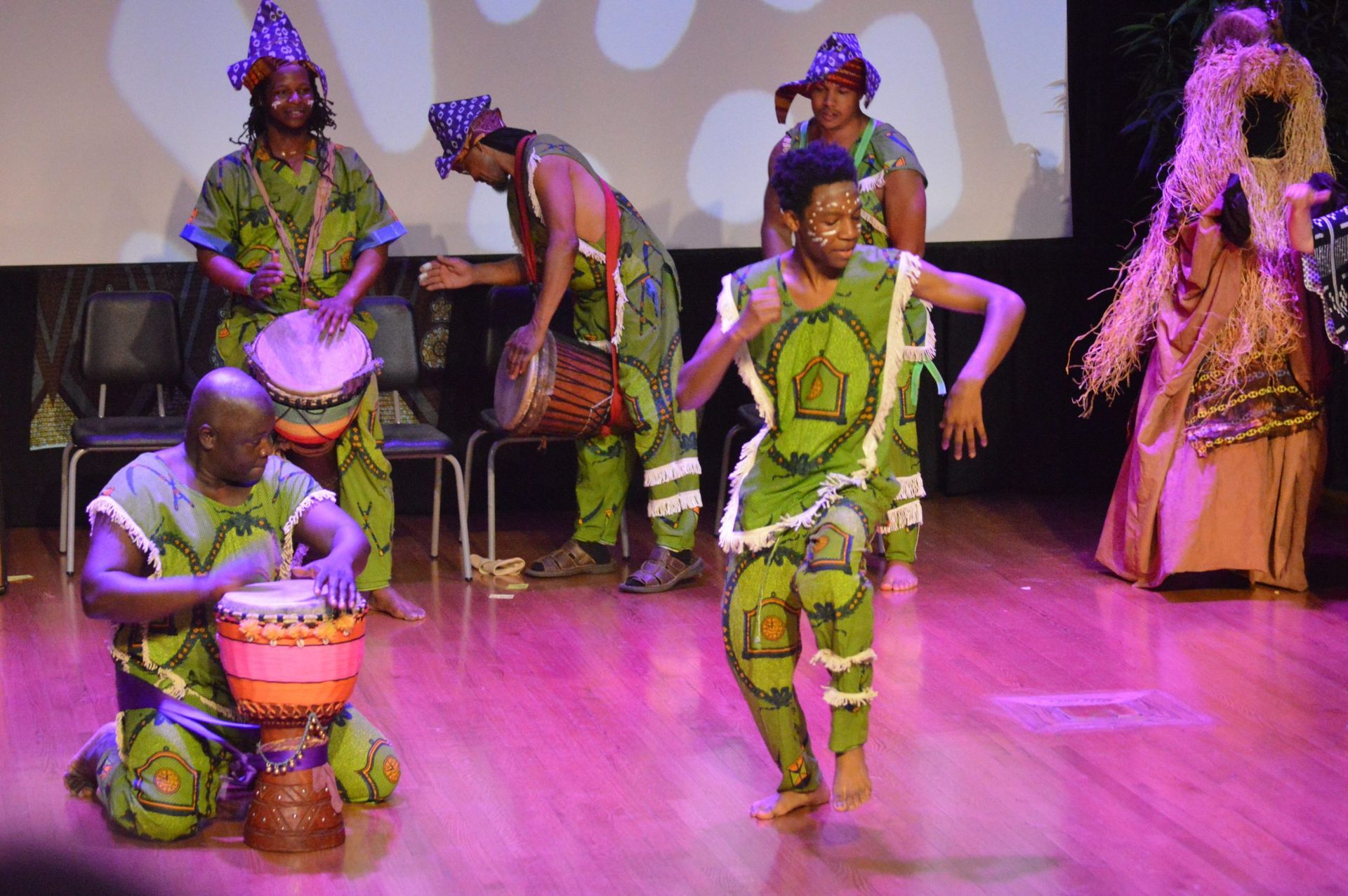 People in green patterned outfits perform with drums and dance on a stage with pink lighting.