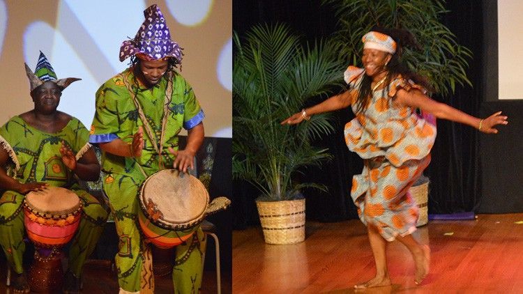 African dancers in colorful costumes perform on stage with drums and palm tree.