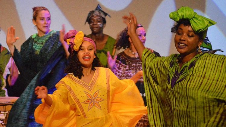 Group of women in colorful African attire, performing on stage, hands raised, joyful expressions.