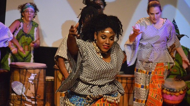 African dancers in colorful traditional dress perform on stage with drums.