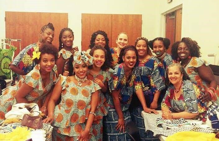 Group of diverse women in colorful African-print dresses, smiling and posing indoors.
