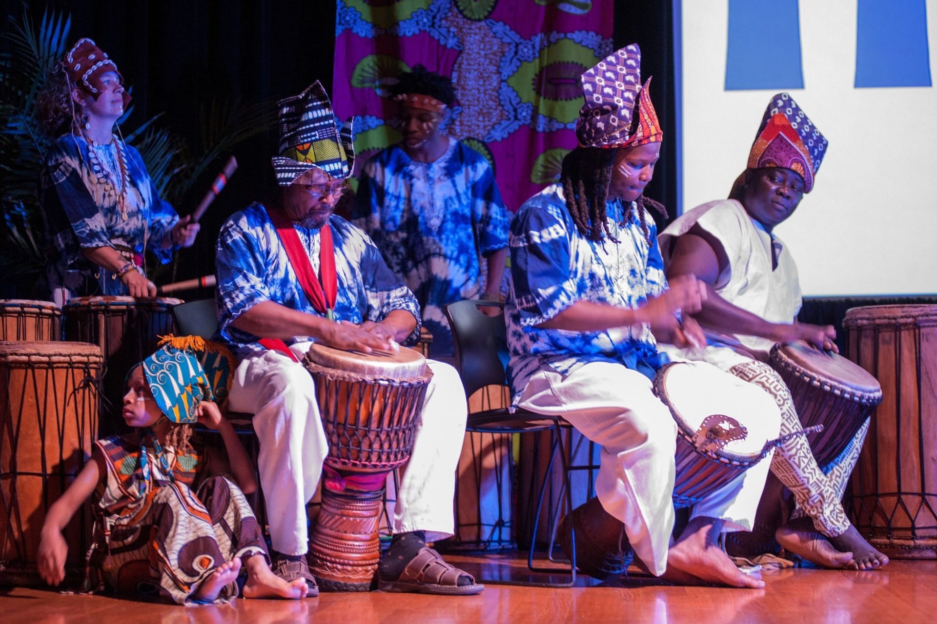 A group of people playing drums onstage, wearing African-style clothing with colorful patterns.