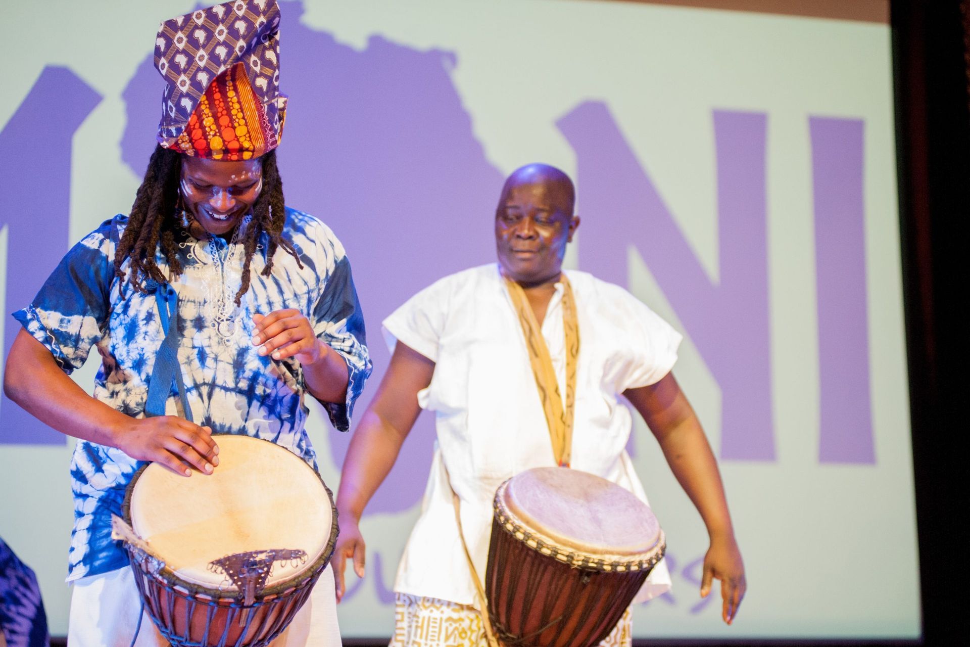 Two men in African attire playing drums onstage. One wears a patterned hat, blue tie-dye shirt.