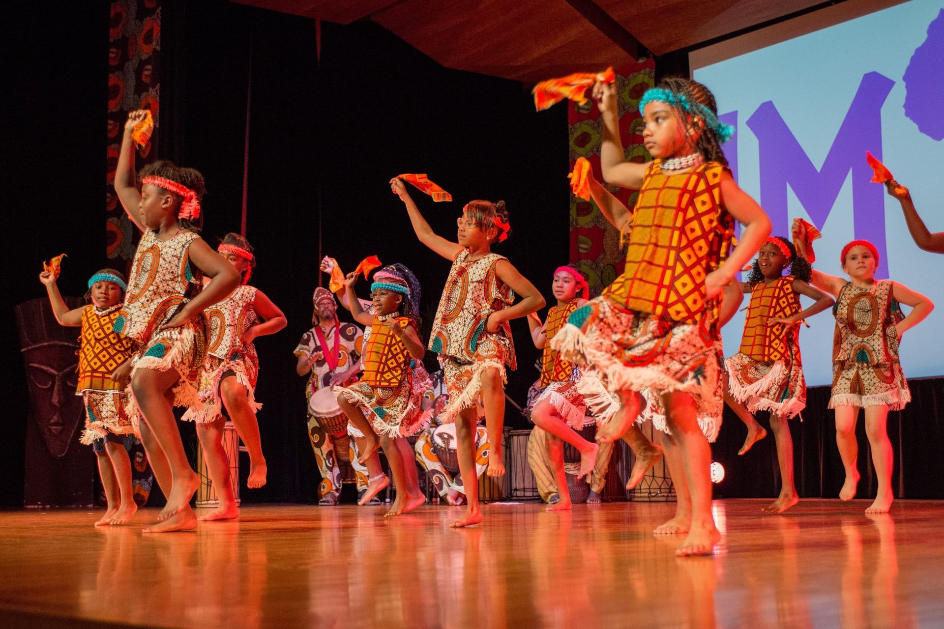 Children in colorful patterned costumes perform a dance on stage, holding orange cloths.