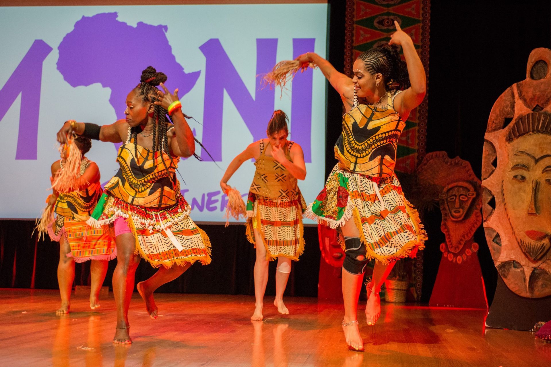 Four dancers in African attire perform onstage; purple backdrop, masks.