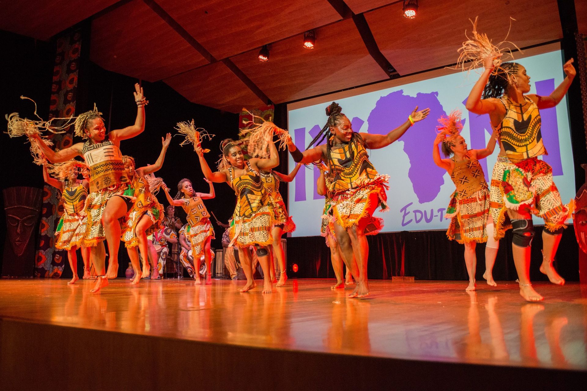 African dancers on stage, in colorful outfits, with arms raised. Background shows a map of Africa and text.