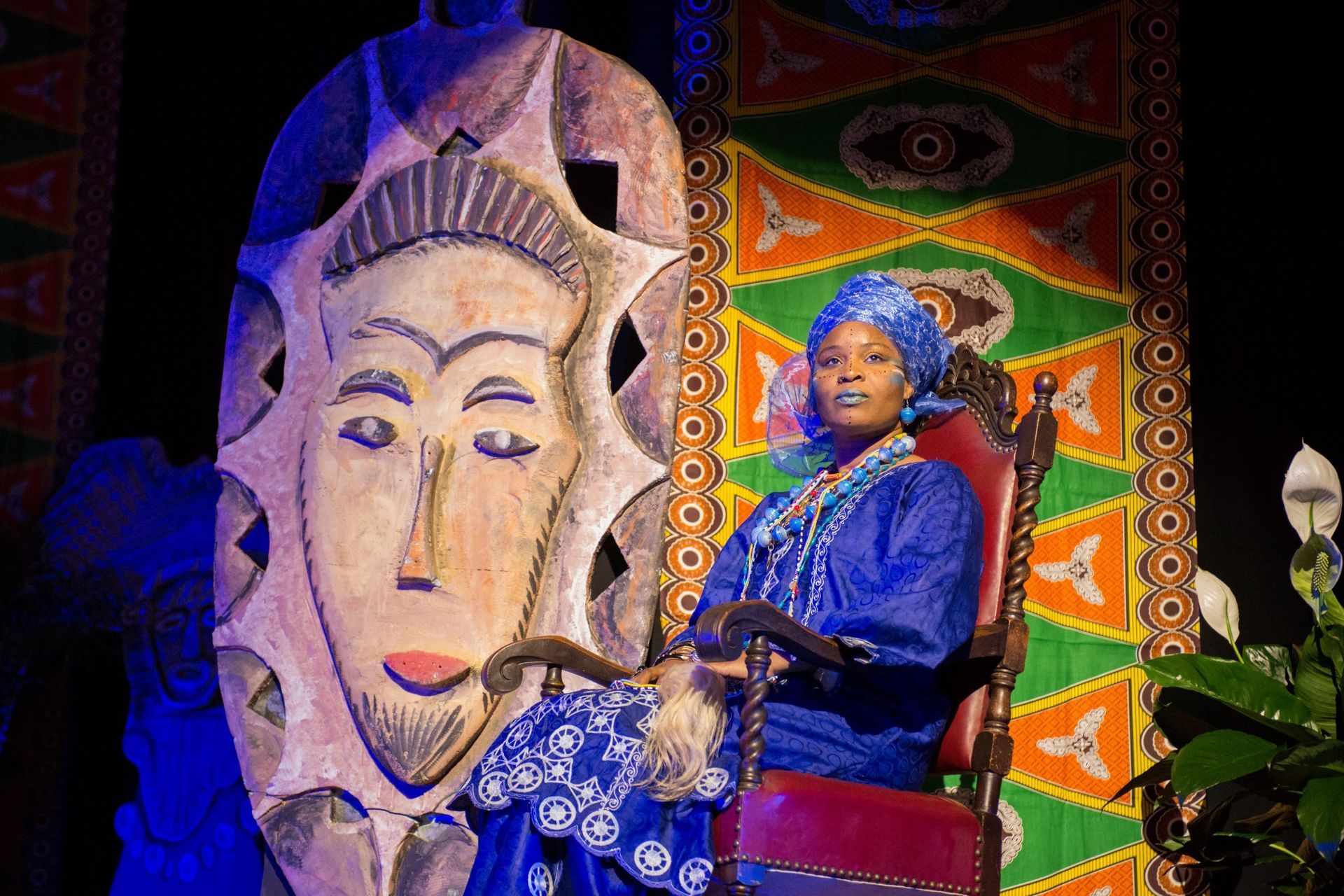 Woman in blue attire sits on a chair. Large mask and colorful textile background.