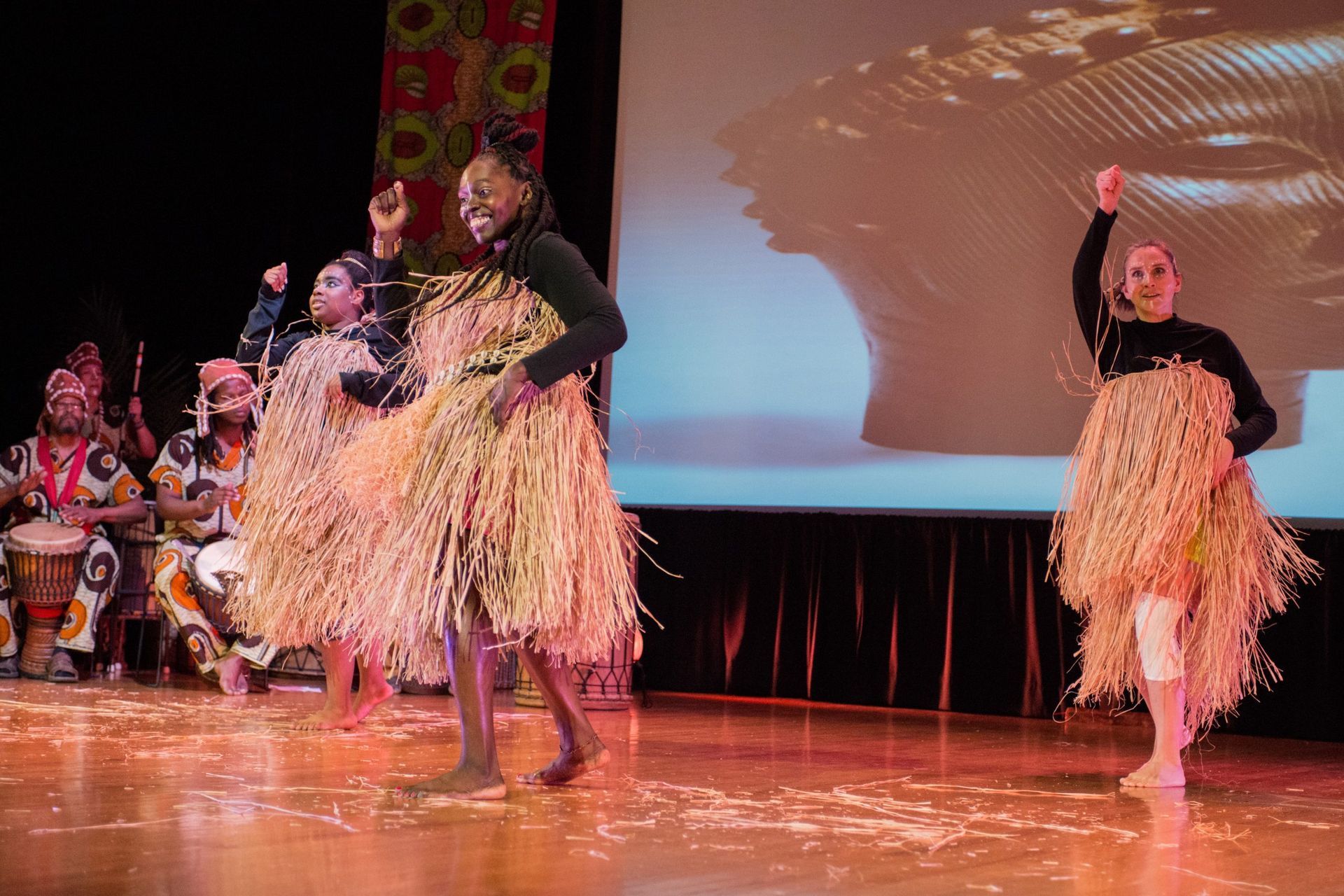 Dancers on stage in straw skirts perform, drummers in the background.