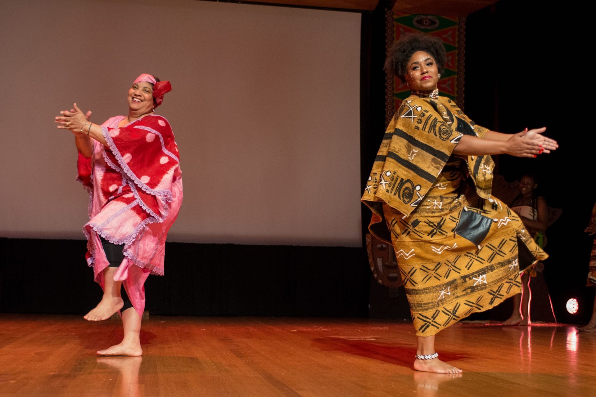 Two women dancing on a wooden stage in colorful traditional African attire.
