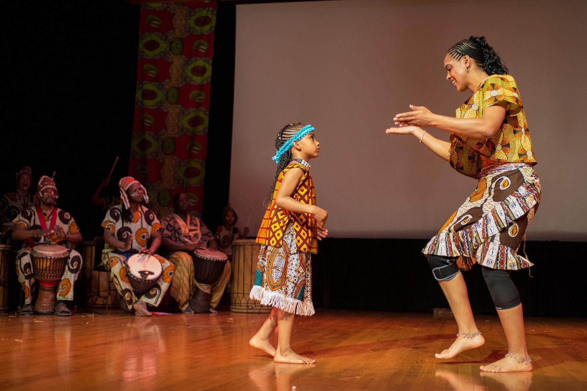 Woman and girl dancing on stage, accompanied by drummers. They wear colorful African-print clothing.