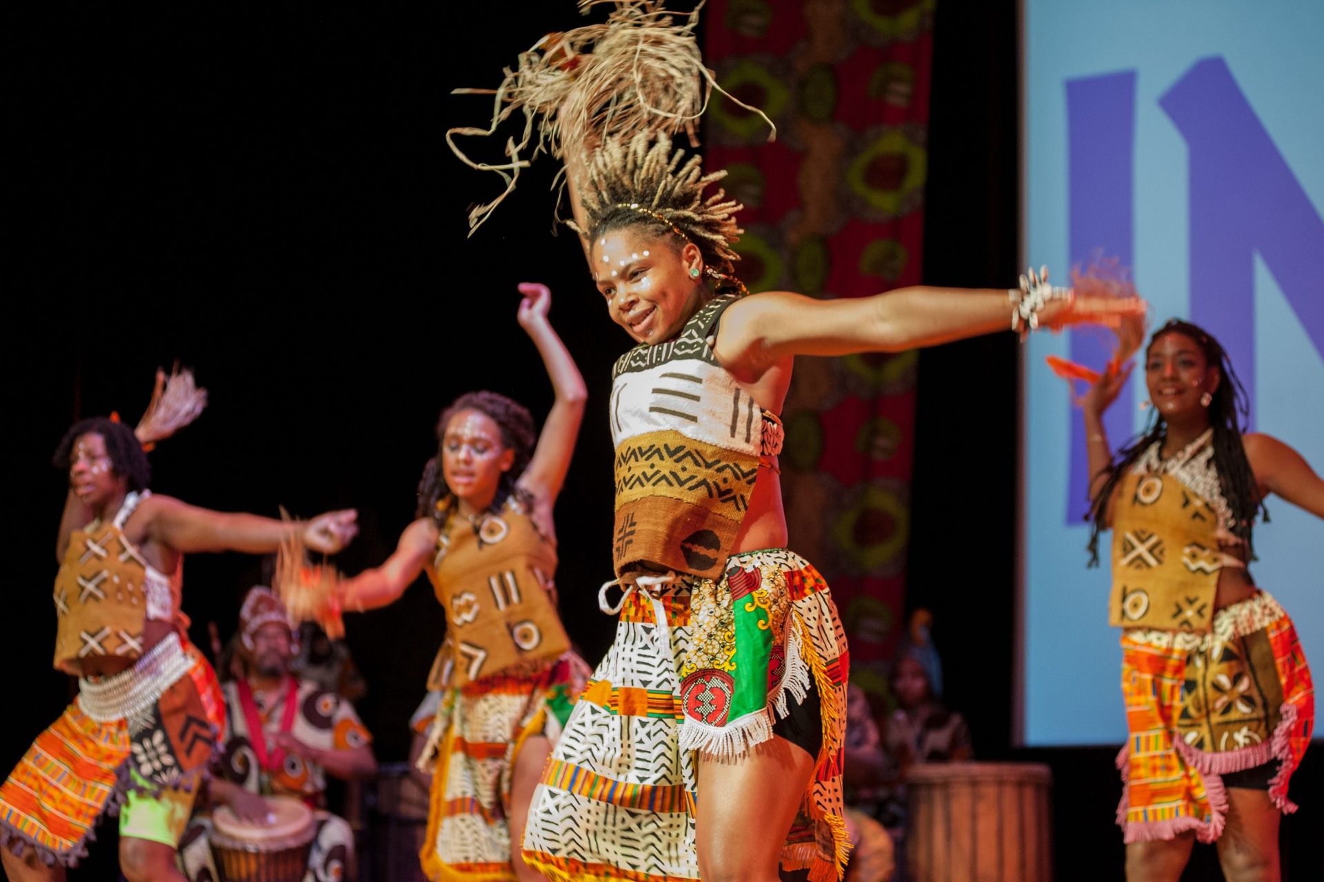 People performing an energetic African dance on stage. They wear colorful, patterned clothing and are smiling.