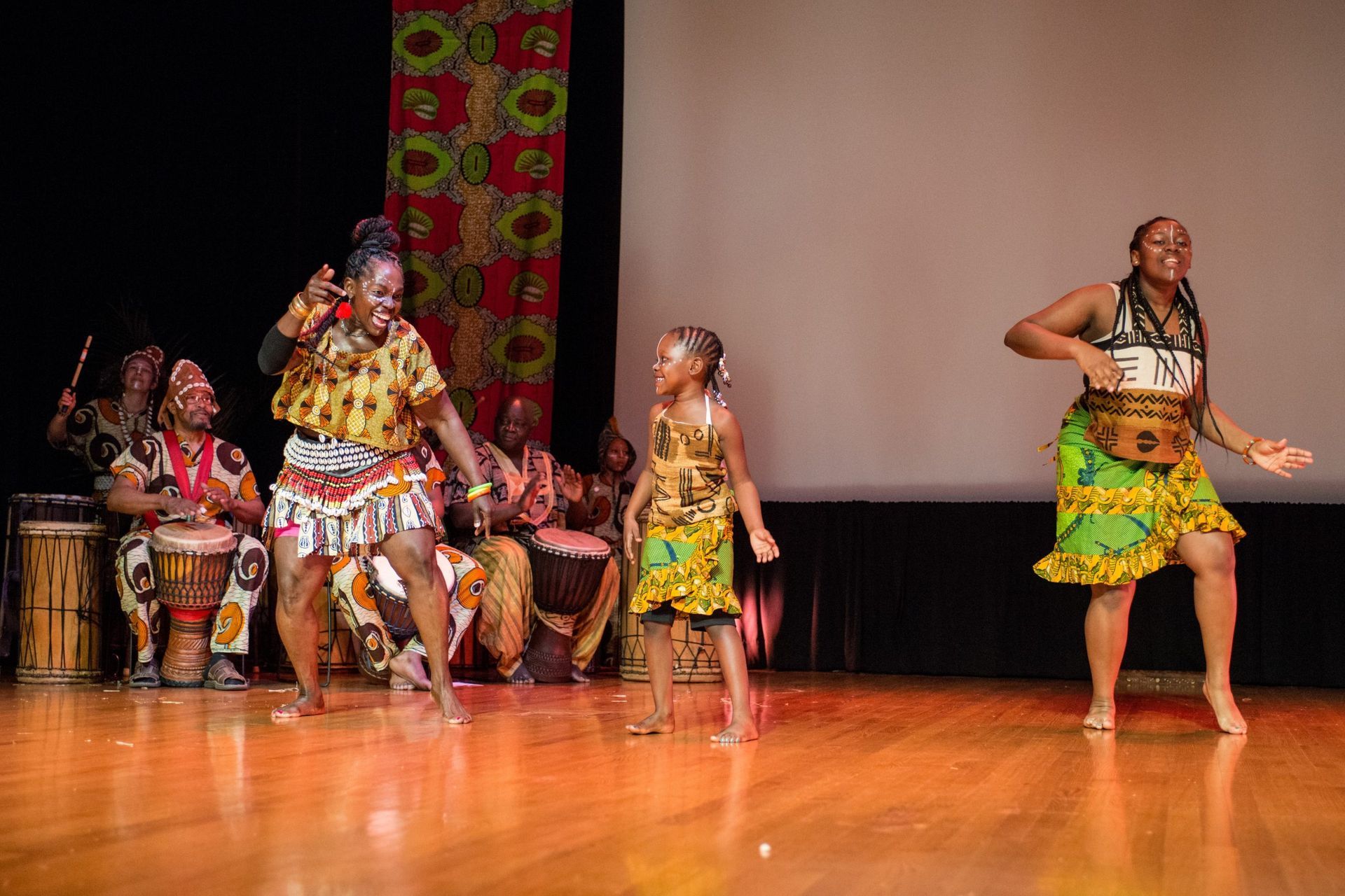 African dancers in colorful outfits perform on stage with drummers in the background.