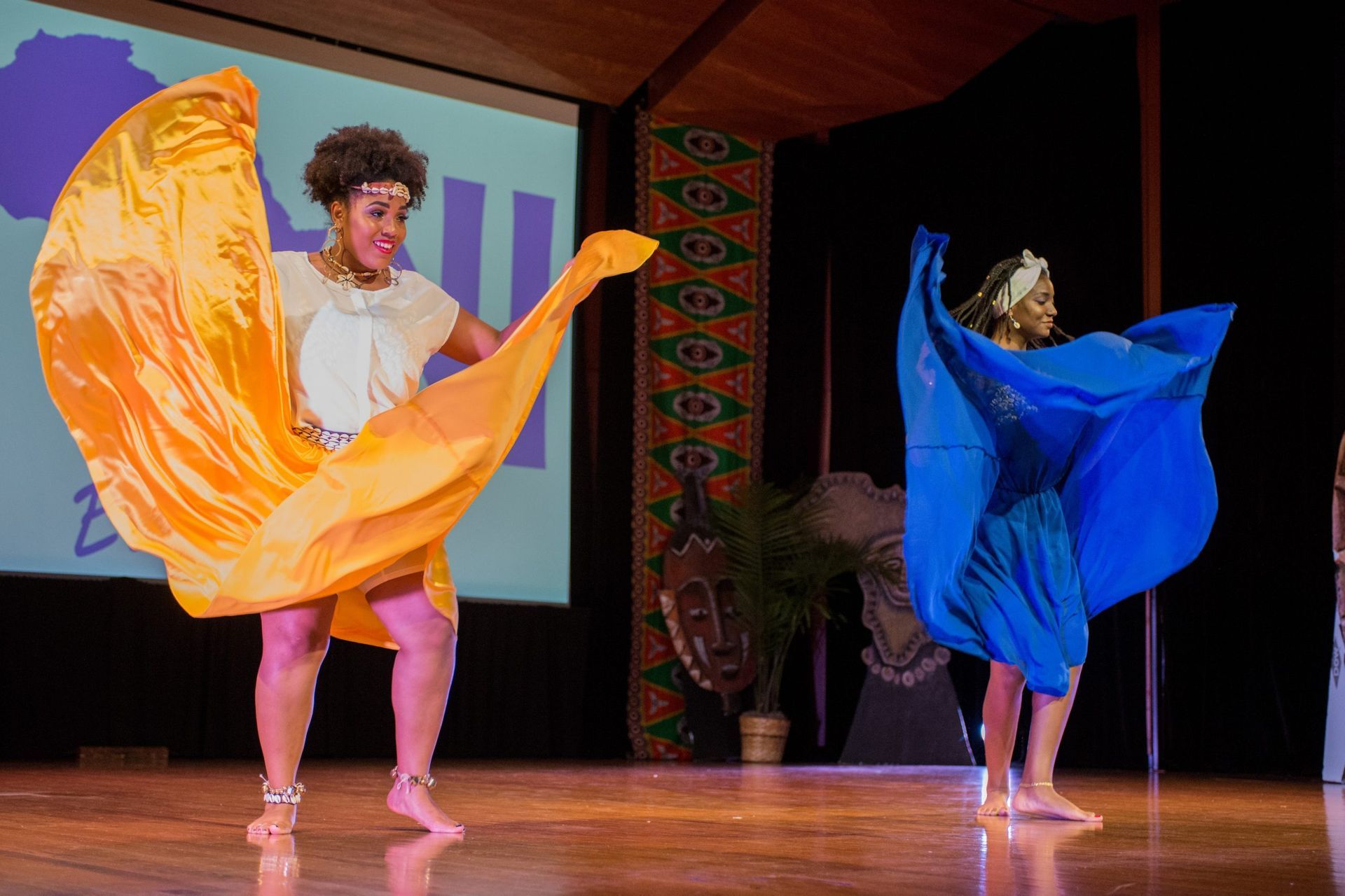 Two dancers on stage, one with orange fabric, the other with blue. They are smiling. Stage setting.