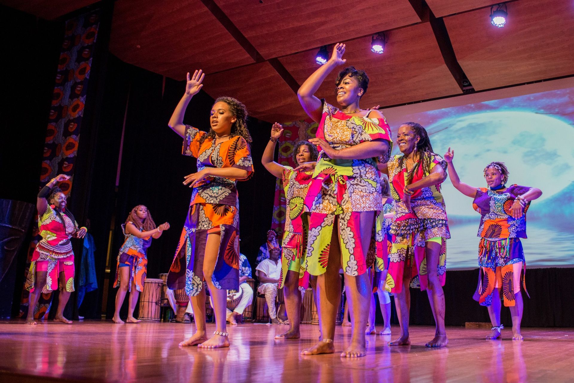 Group of people in vibrant African-print outfits dance on a stage, lit with stage lights and a backdrop of a moon.