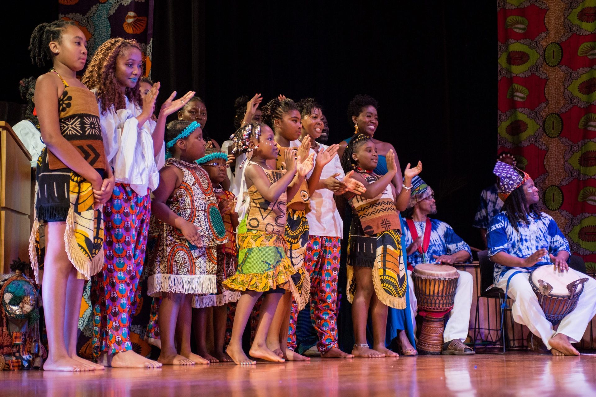 Children in colorful African attire clap on a stage with musicians.