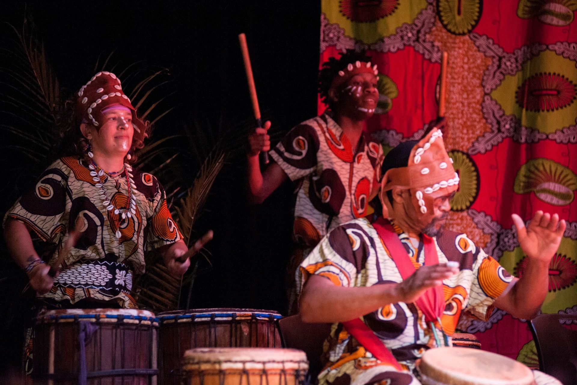 Group of African drummers in colorful patterned shirts and headdresses playing drums onstage.