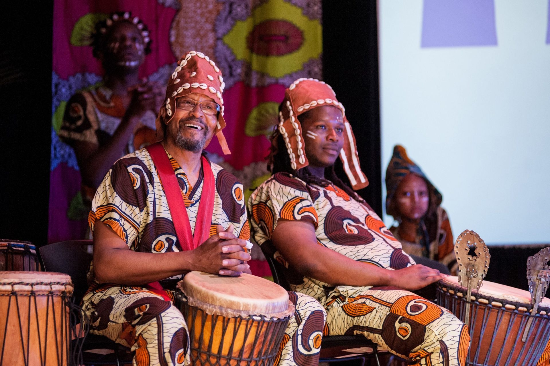 People in African-print outfits playing drums onstage; audience in background, colorful backdrop.