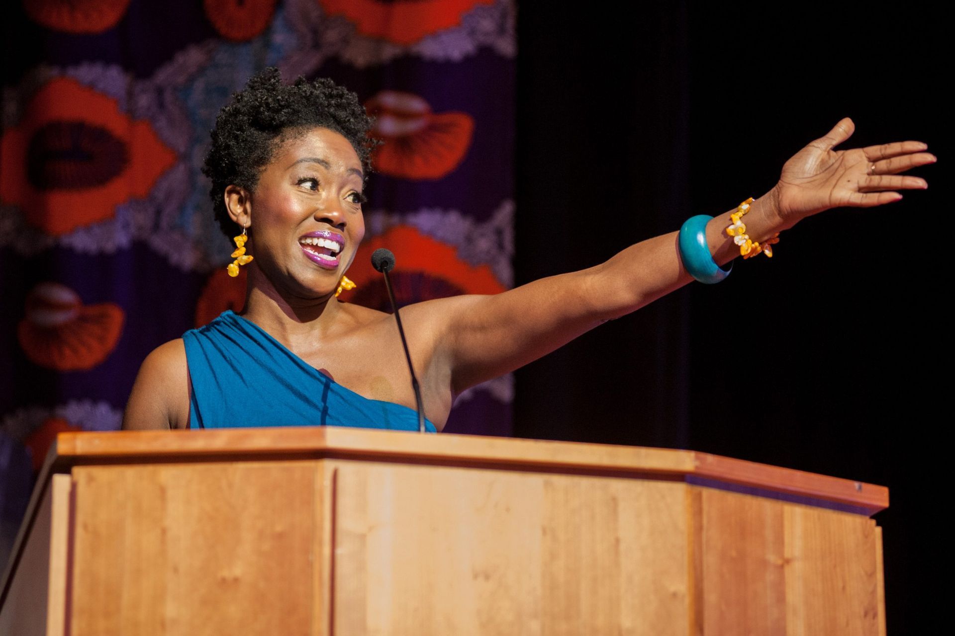 Woman giving a speech at a podium, smiling, gesturing with right arm. Teal dress, gold and turquoise jewelry.
