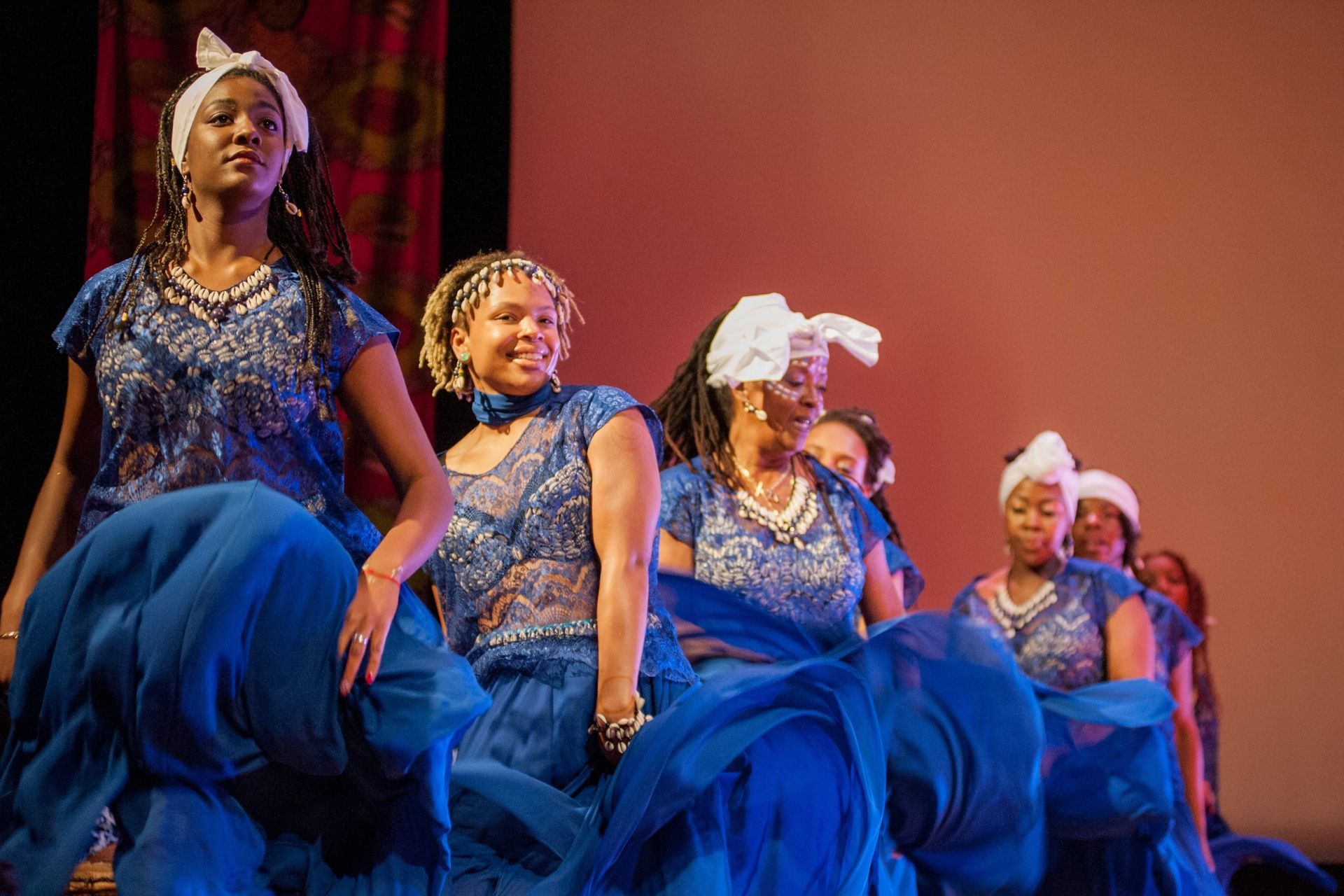 Women in blue dresses and headscarves dance on stage with red backdrop.