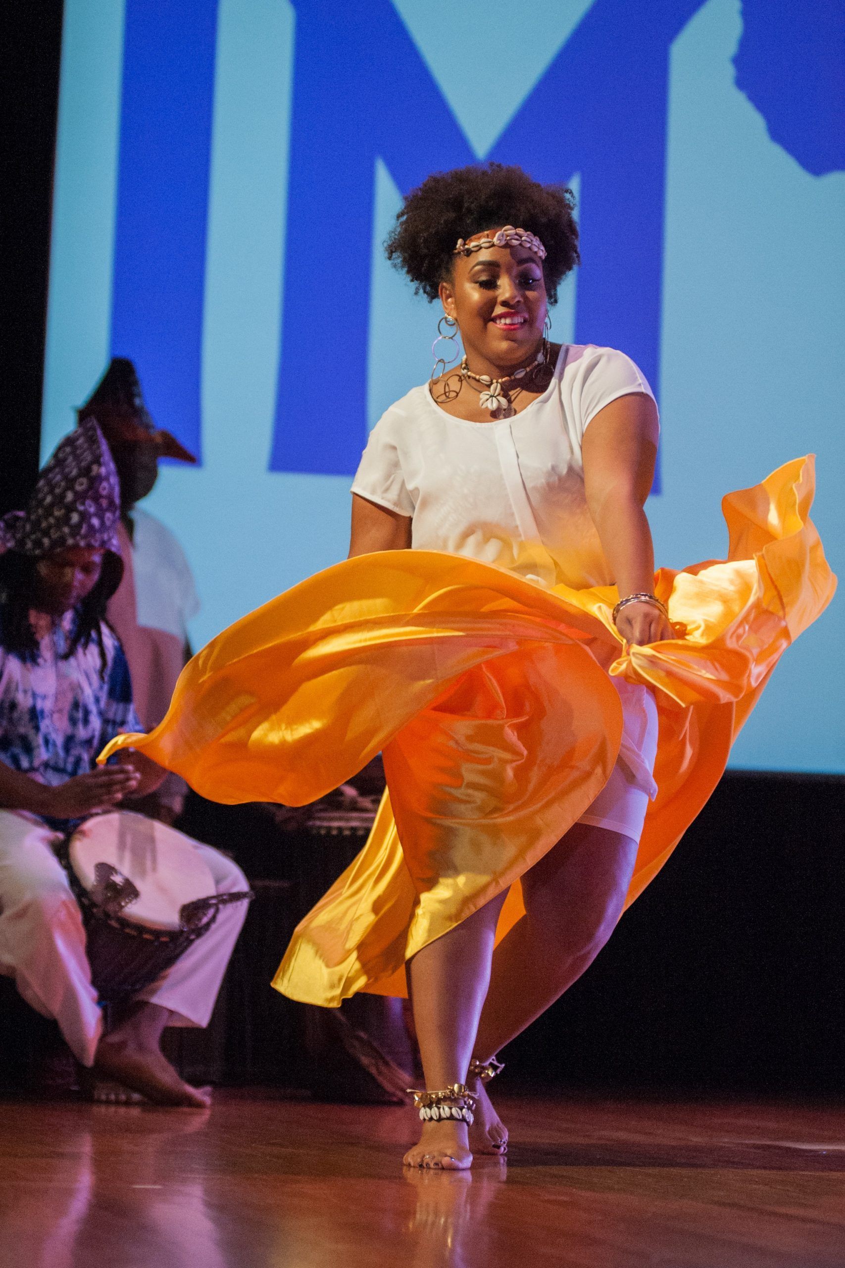 Woman in white top and orange skirt dancing on stage, smiling.
