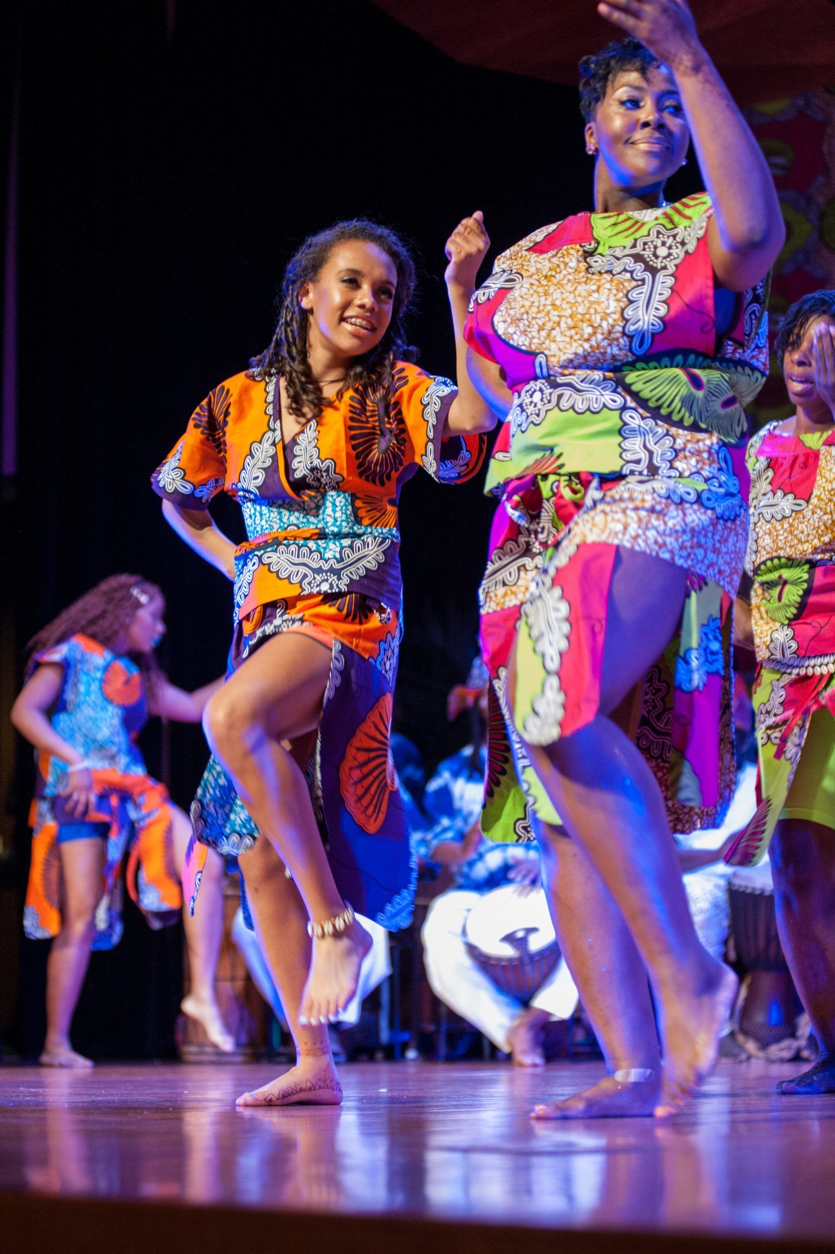 Dancers in vibrant African print dresses perform on stage. Barefoot, smiling, they move expressively.