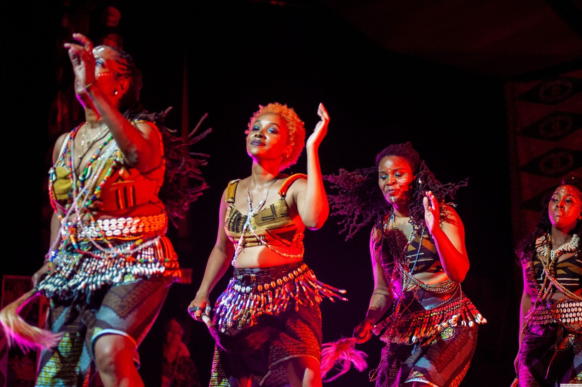 Women in vibrant costumes dance on stage, arms raised under stage lighting.