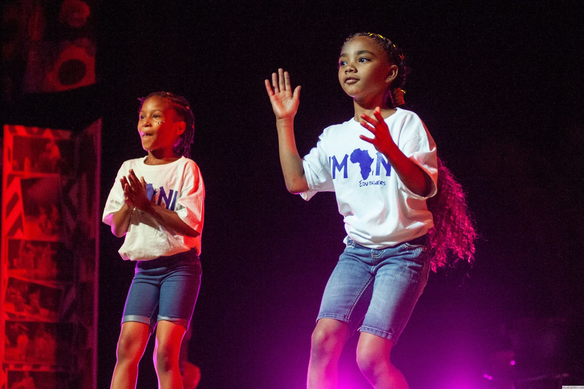 Two young girls in white shirts and denim shorts dance on stage.