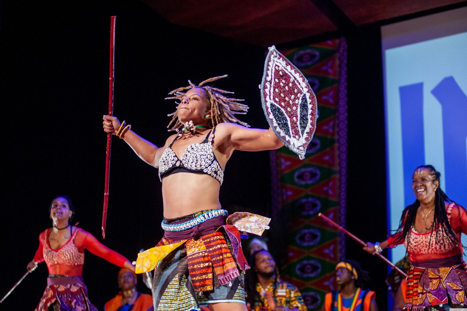 African dancers perform on stage; woman holds shield and spear, wearing a patterned top and skirt.