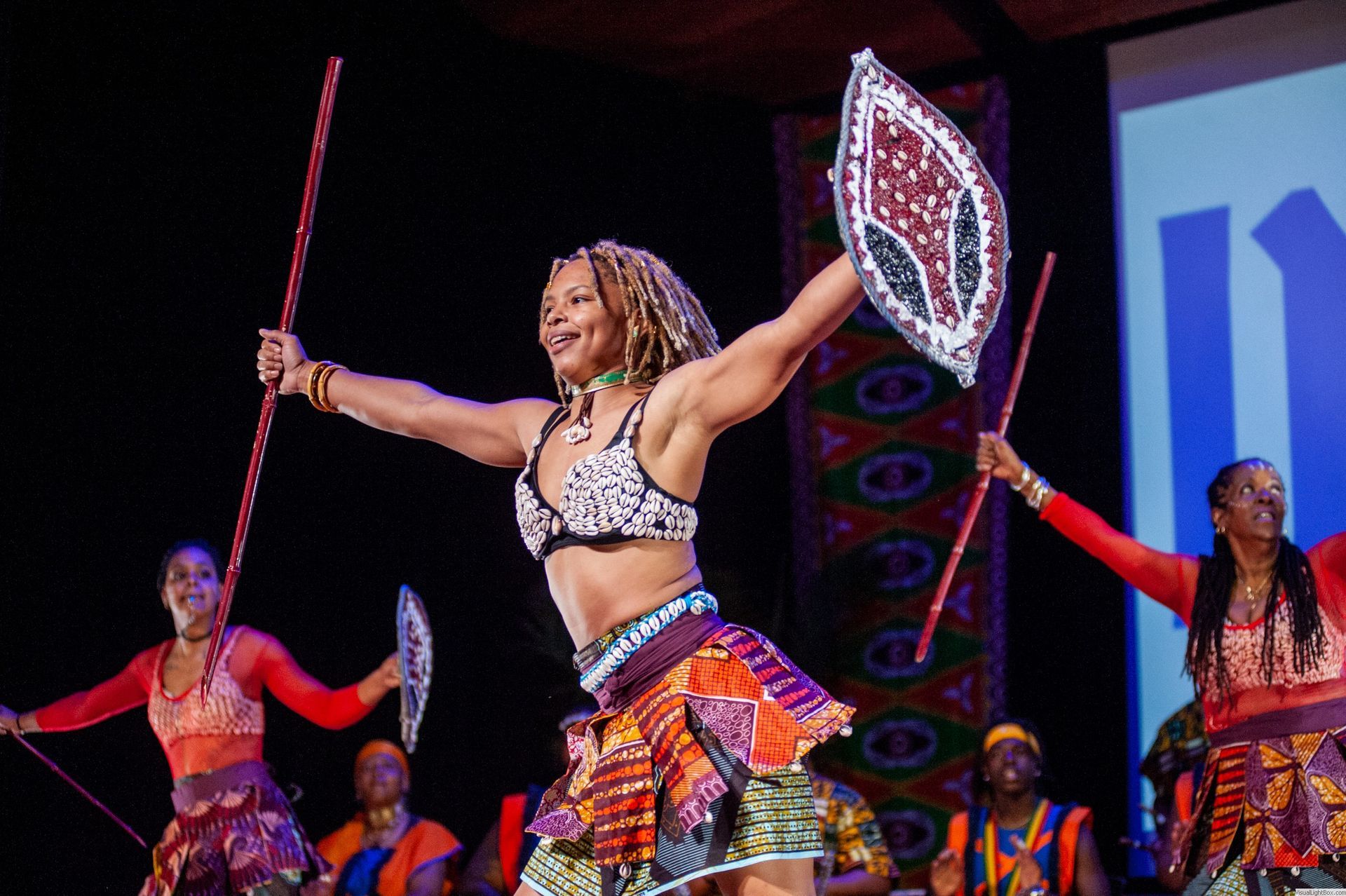 African dancers in colorful costumes perform on stage, holding shields and sticks, arms raised.