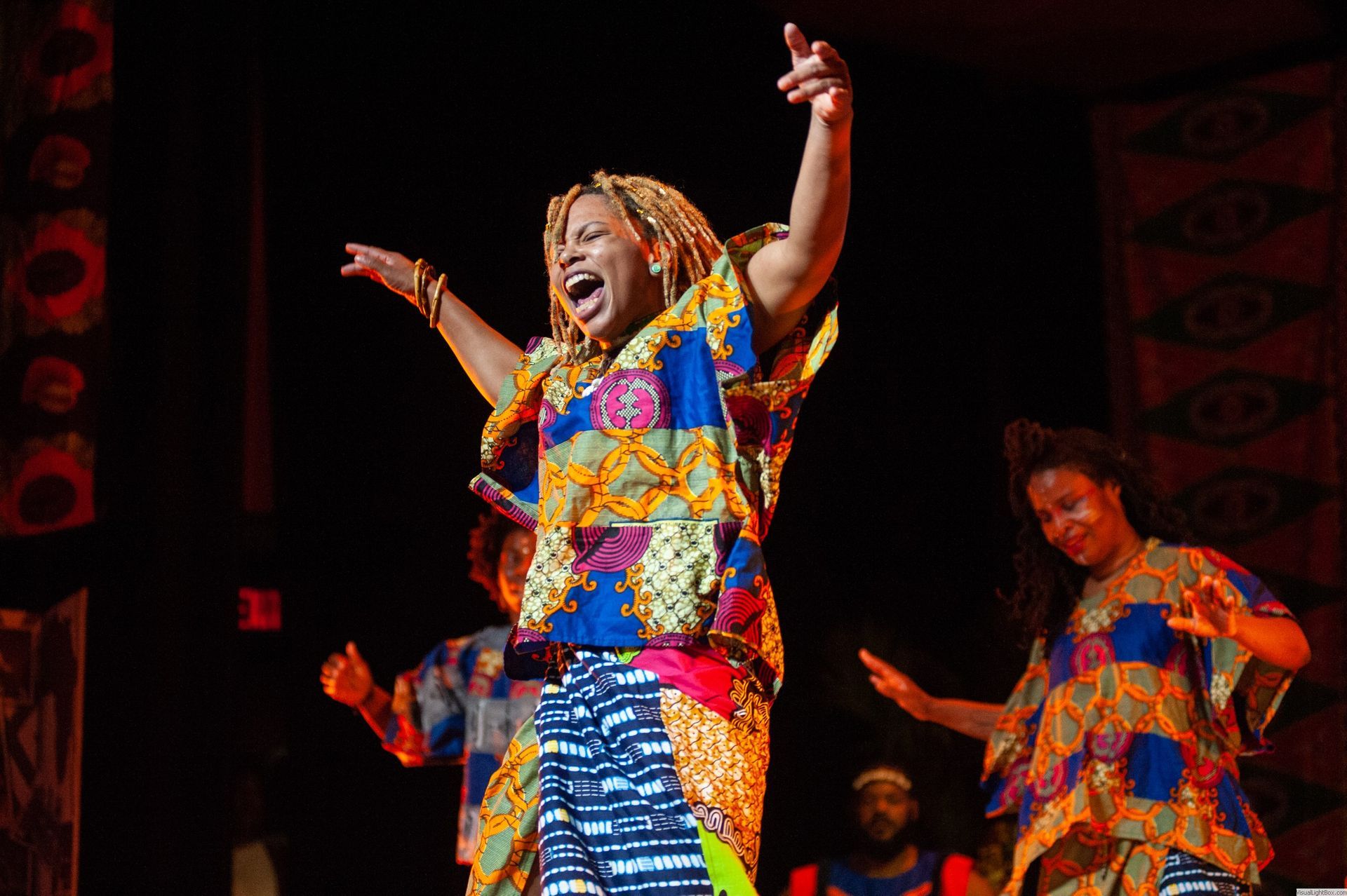 Woman in colorful African print dress singing and dancing on stage, arms raised.