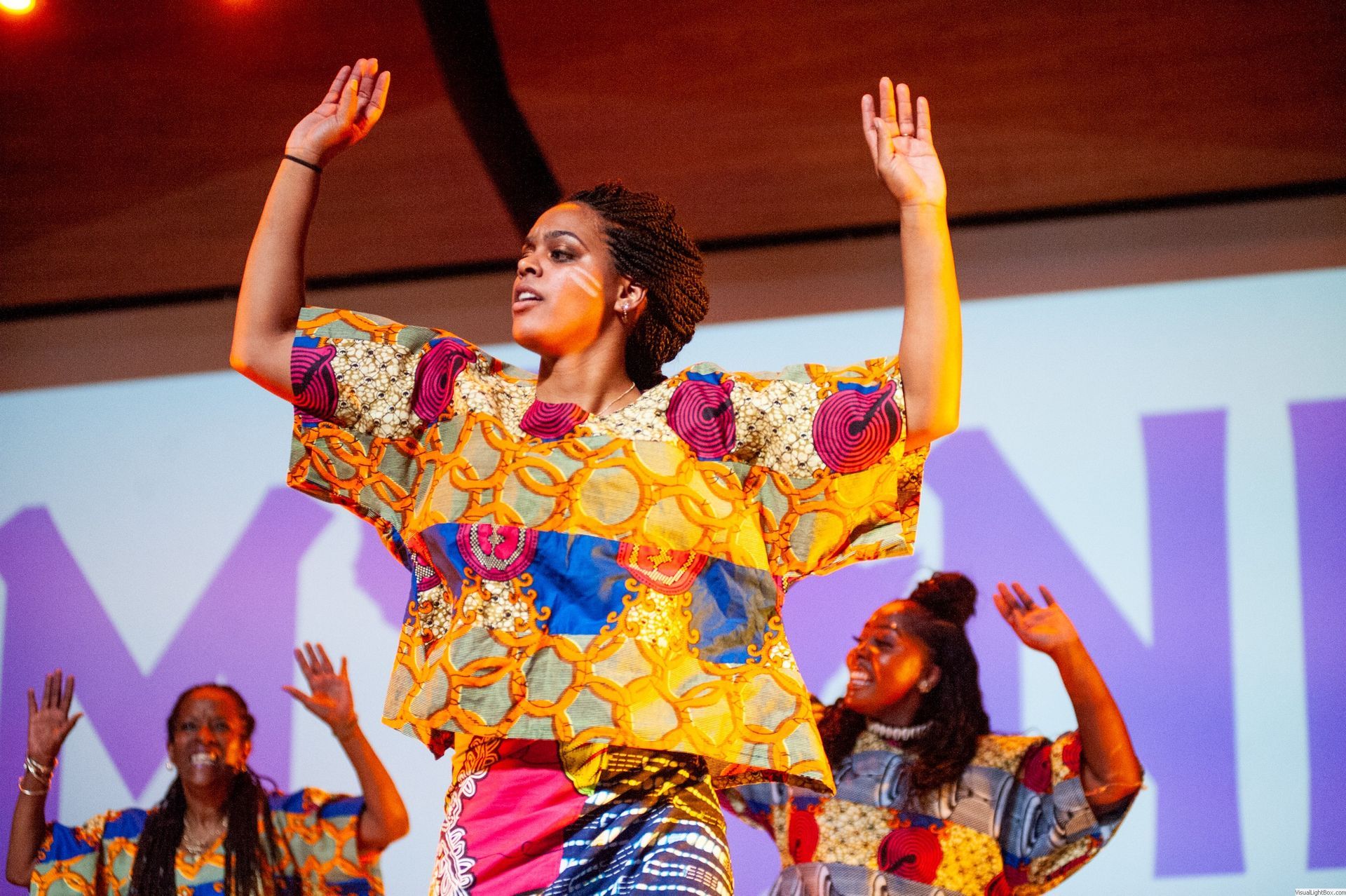 Dancers in colorful African print outfits perform on stage with arms raised.