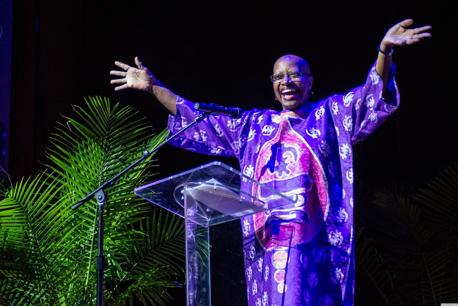Woman with arms raised, wearing a purple patterned robe, speaking at a podium, stage with plants.