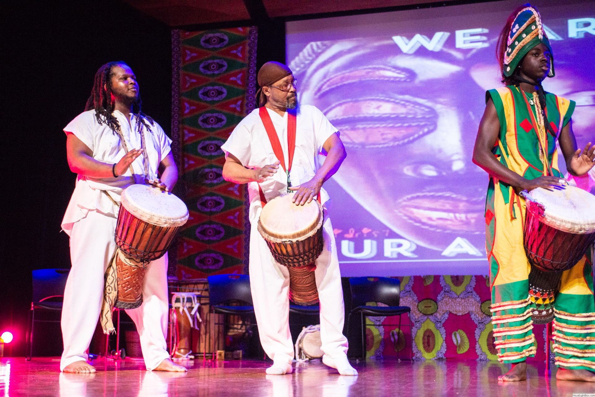 Three men in white clothing play African drums on stage, with a projected face behind them.