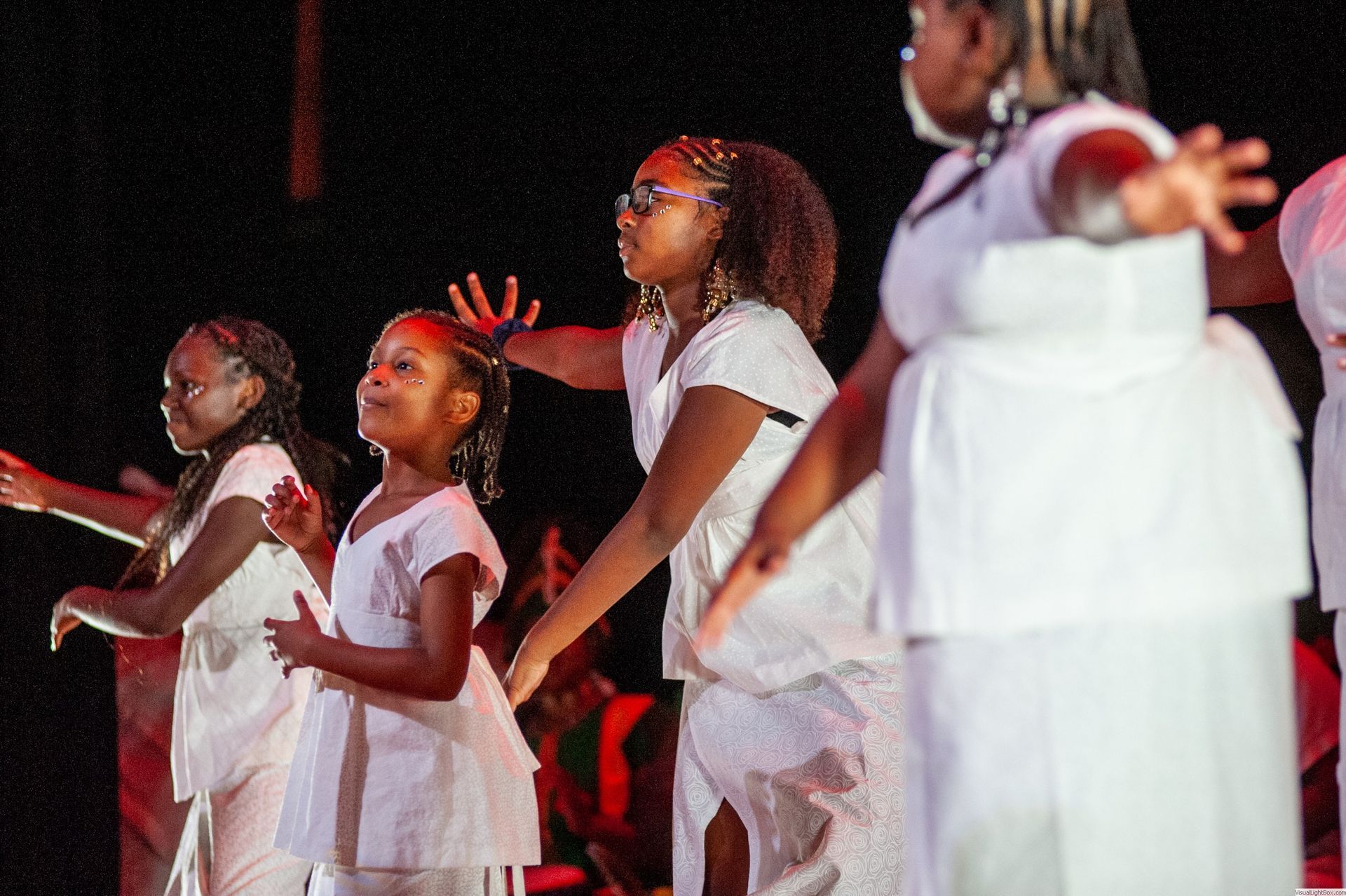 Group of Black women and girls in white outfits performing on a stage with outstretched arms.