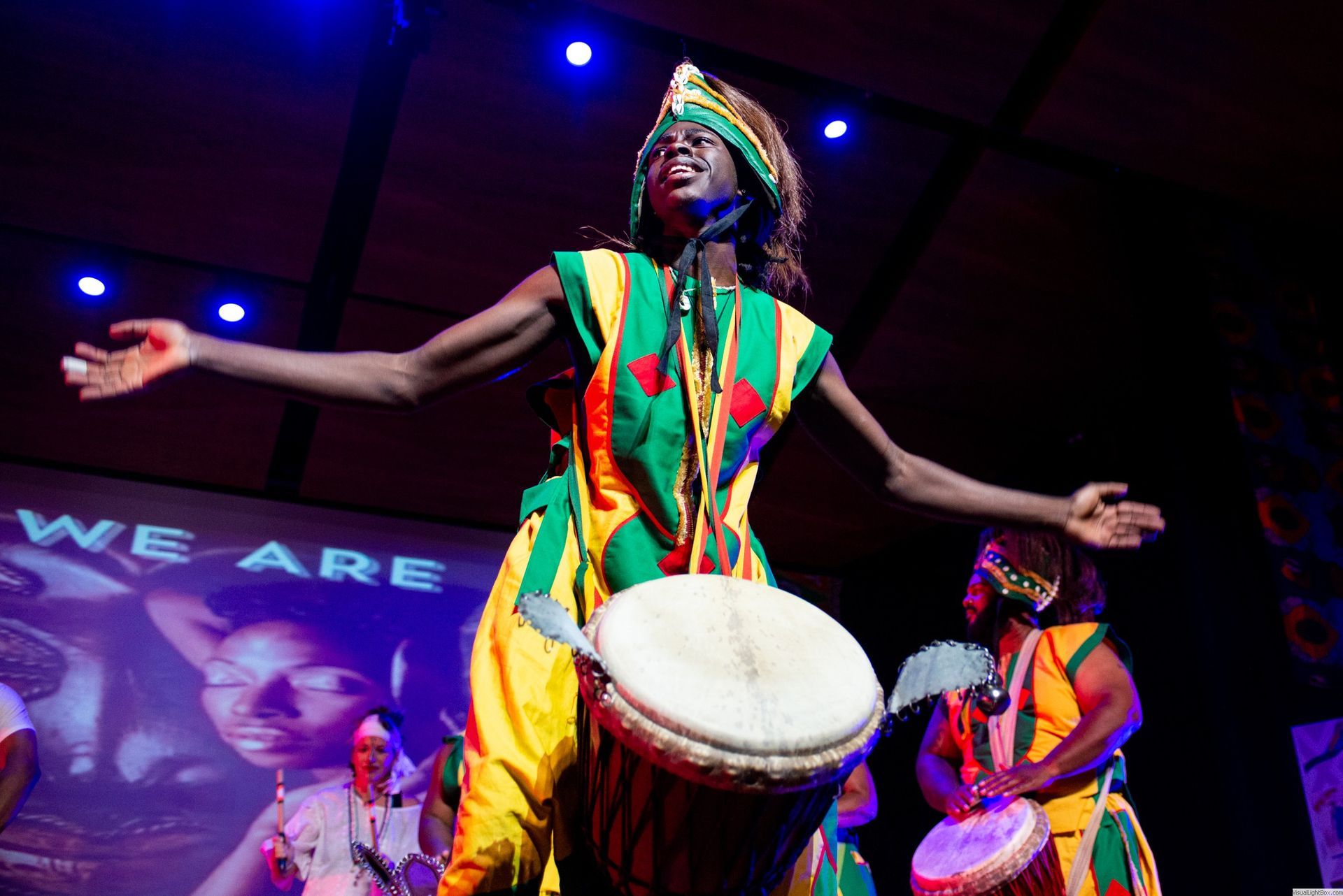 Man in colorful African garb playing drum, stage performance, arms outstretched.