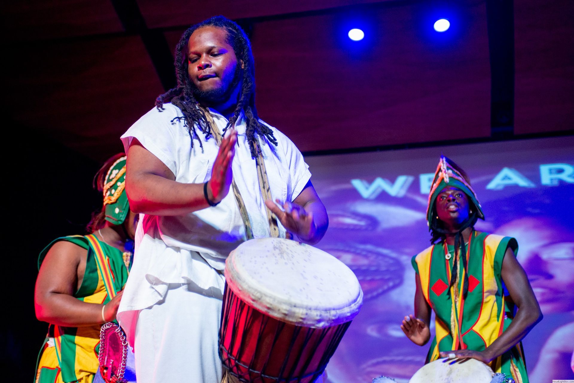 Man in white playing drum onstage with two others in colorful outfits.