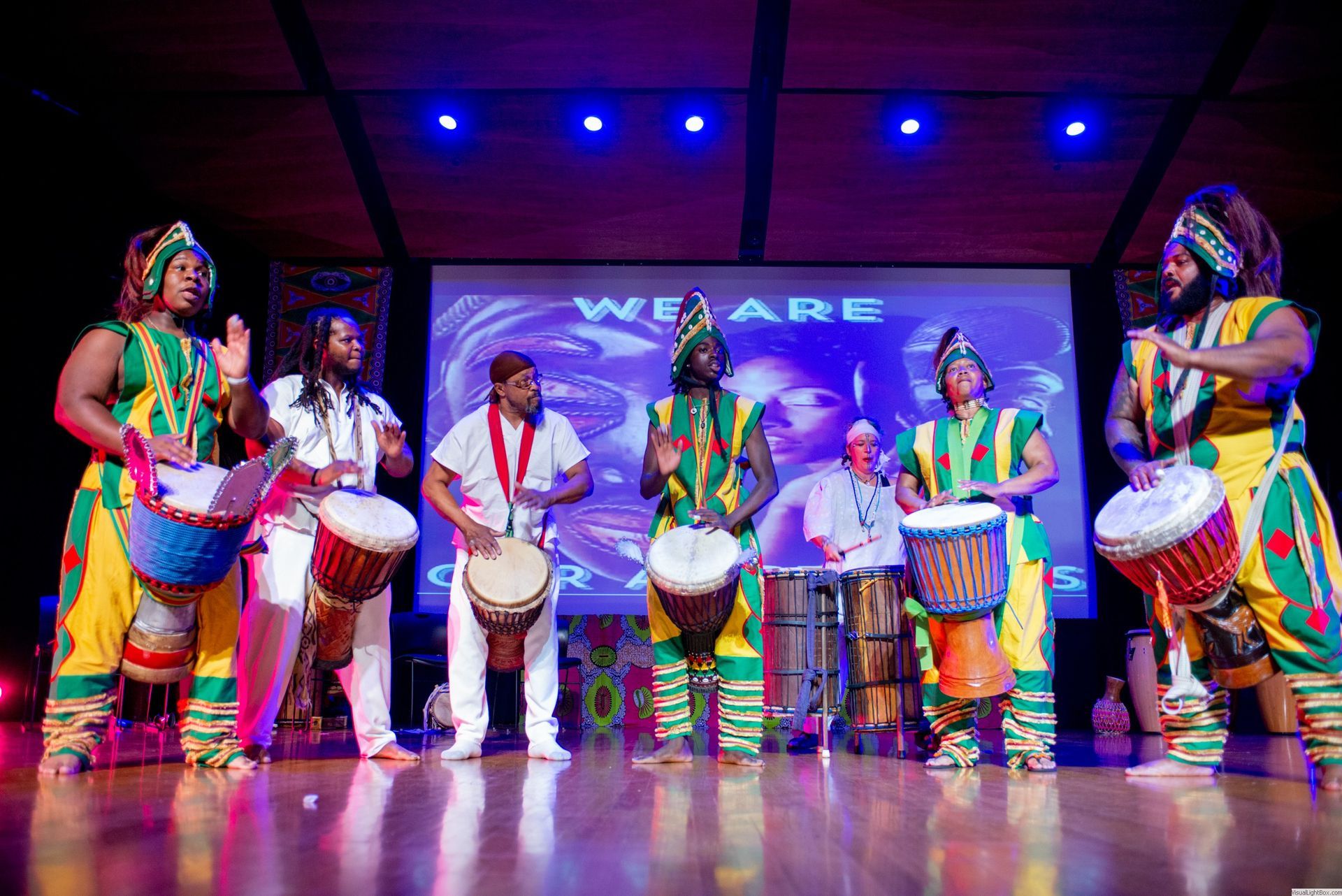 A group of people in colorful African outfits playing drums on stage with a screen that says 